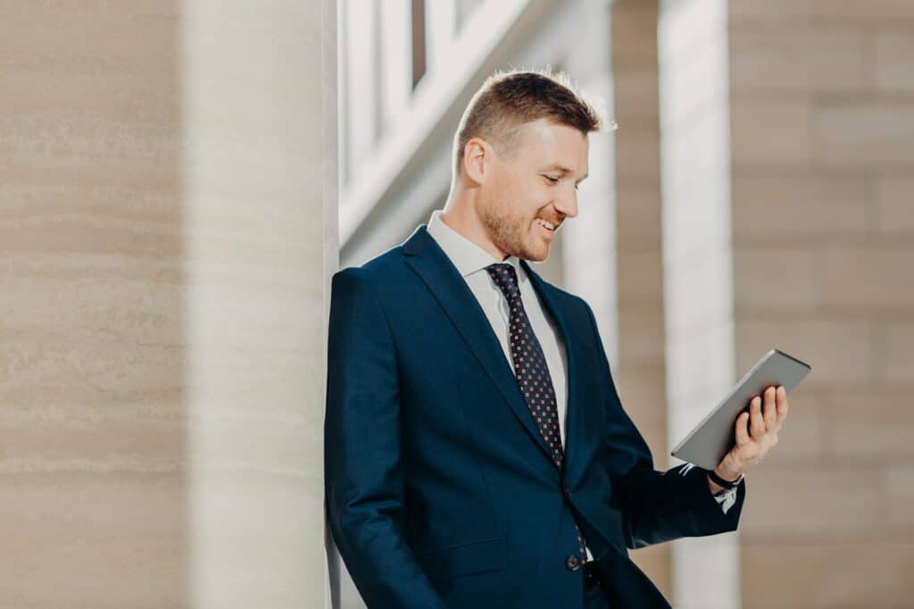 Businessman in formal suit, holds digital tablet in hands and reads business news on website